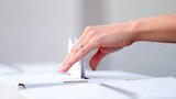 Eine Hand, die einen Zettel in eine Wahlurne steckt.